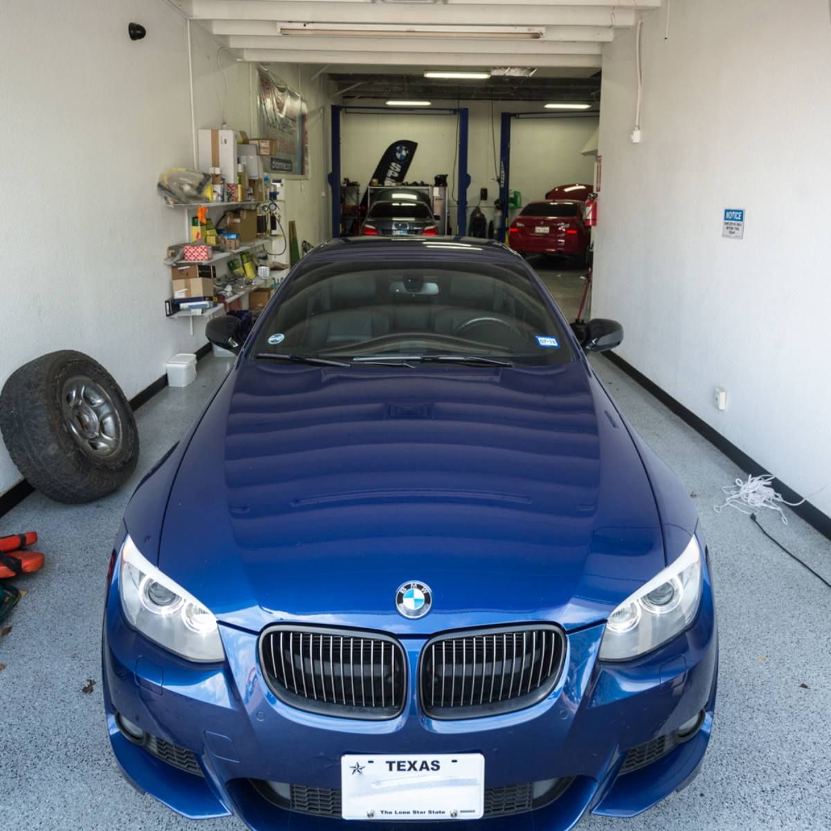 Blue BMW in garage; tire and tools in foreground, car on lift in the back.