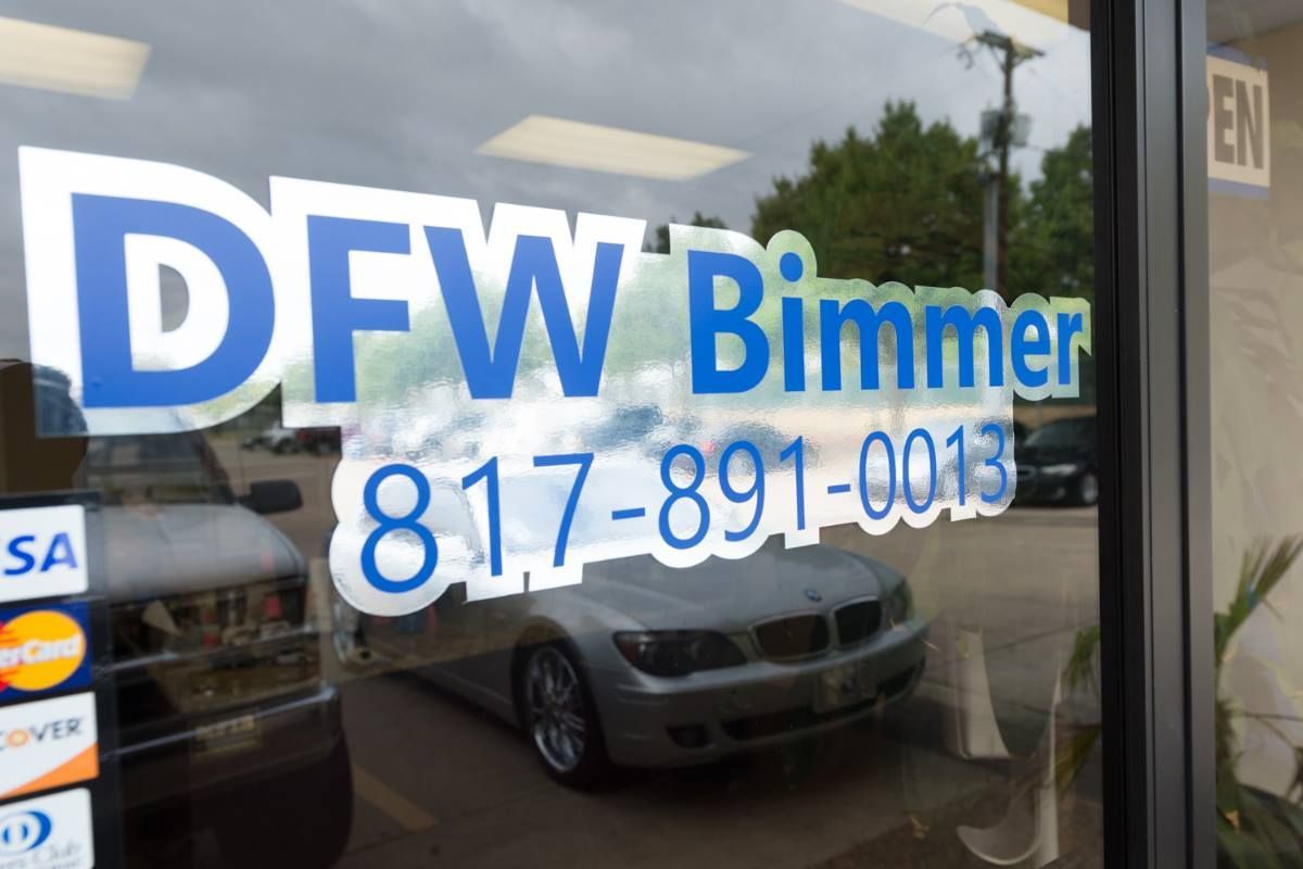 Window sign for DFW Bimmer with phone number. Silver BMW and car parts visible through the glass.