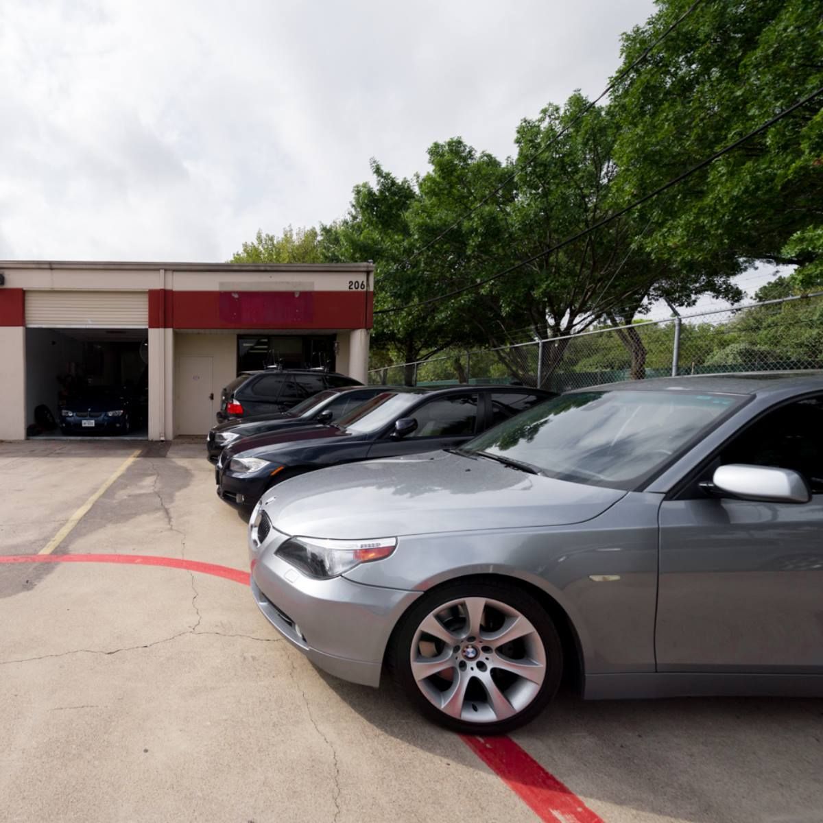 Several cars parked outside a repair shop. A silver car is in the foreground. Trees and a cloudy sky are visible.