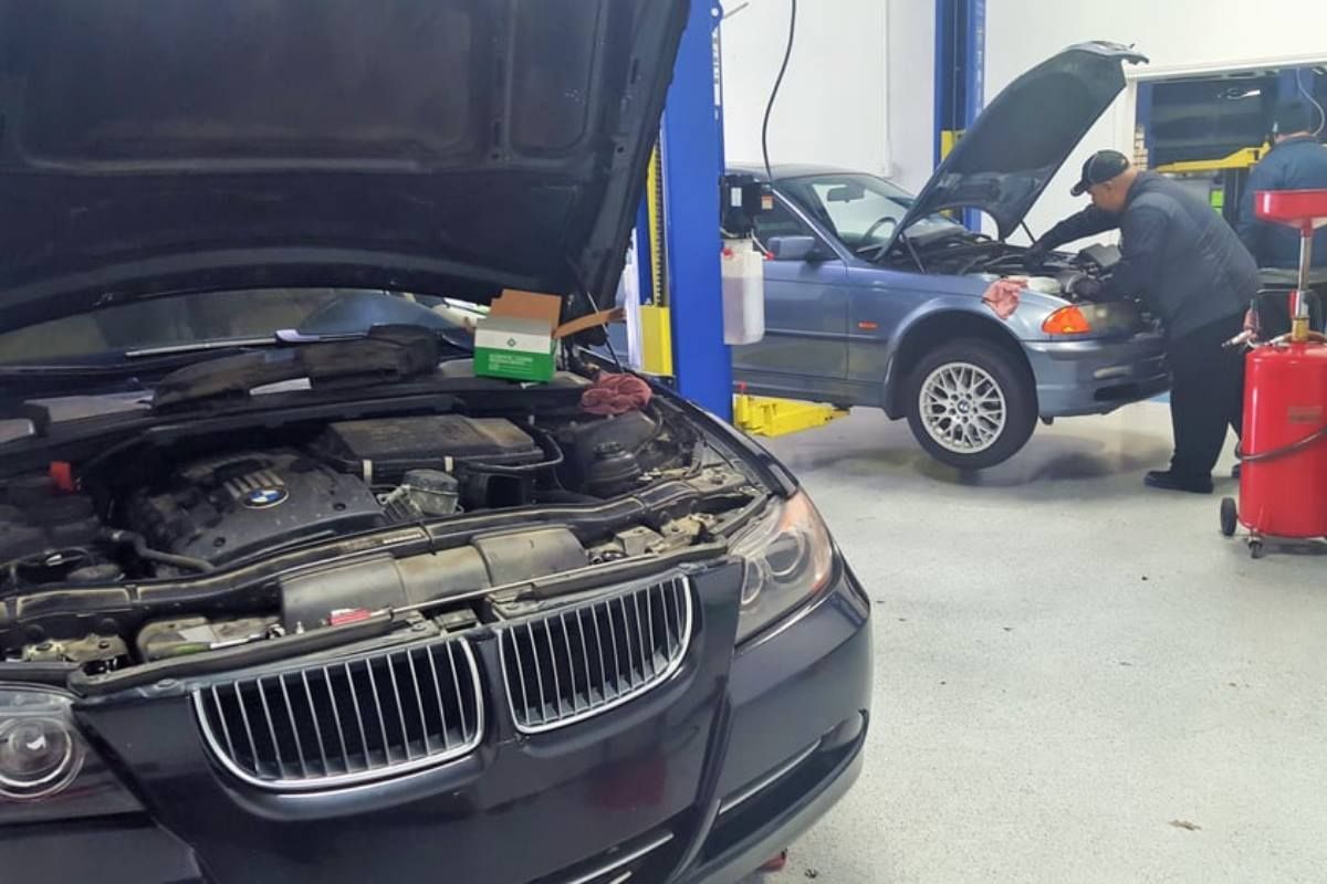 Cars with open hoods in a repair shop; a mechanic works on a lifted blue vehicle.