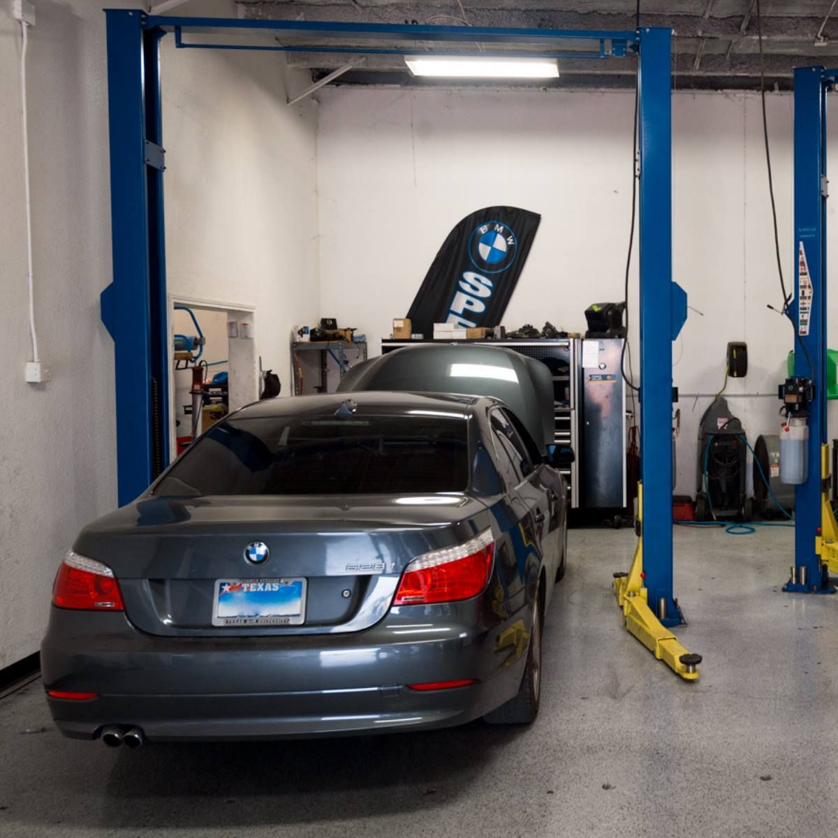 Gray BMW sedan on a lift in a garage with blue posts. The hood is open, and a BMW sign is visible.