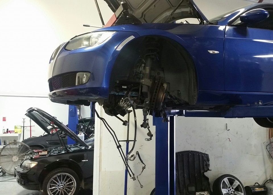 Blue car on a lift in a garage, front wheel removed, hood open, another car in the background.