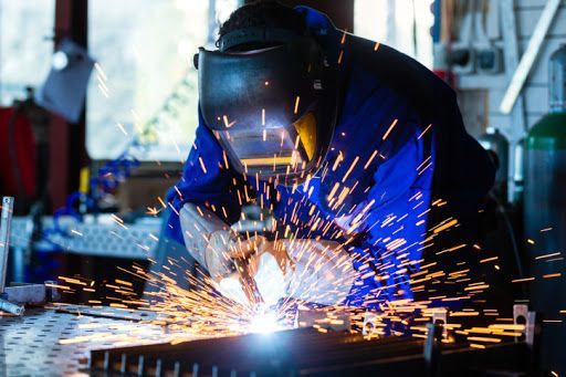 A person wearing a welding helmet and protective blue gear works on metal, producing bright orange sparks in a workshop.