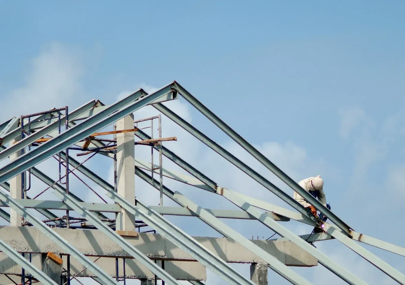 A construction worker in a hard hat secures metal roof beams on an unfinished building against a blue sky.