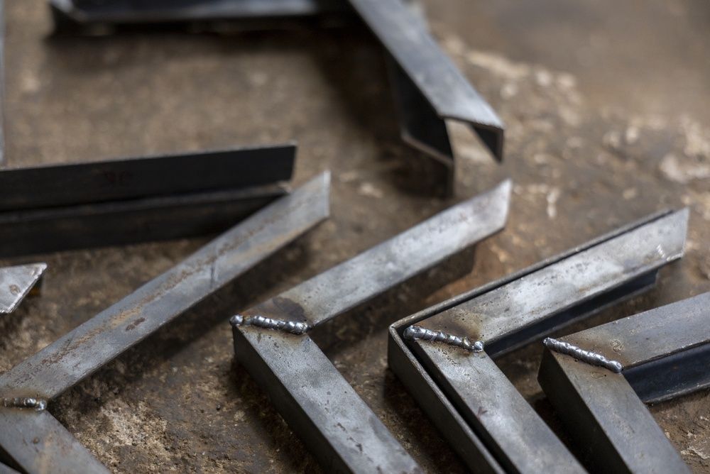 Several cut and welded steel L-shaped bracket components arranged on a textured, workshop surface.