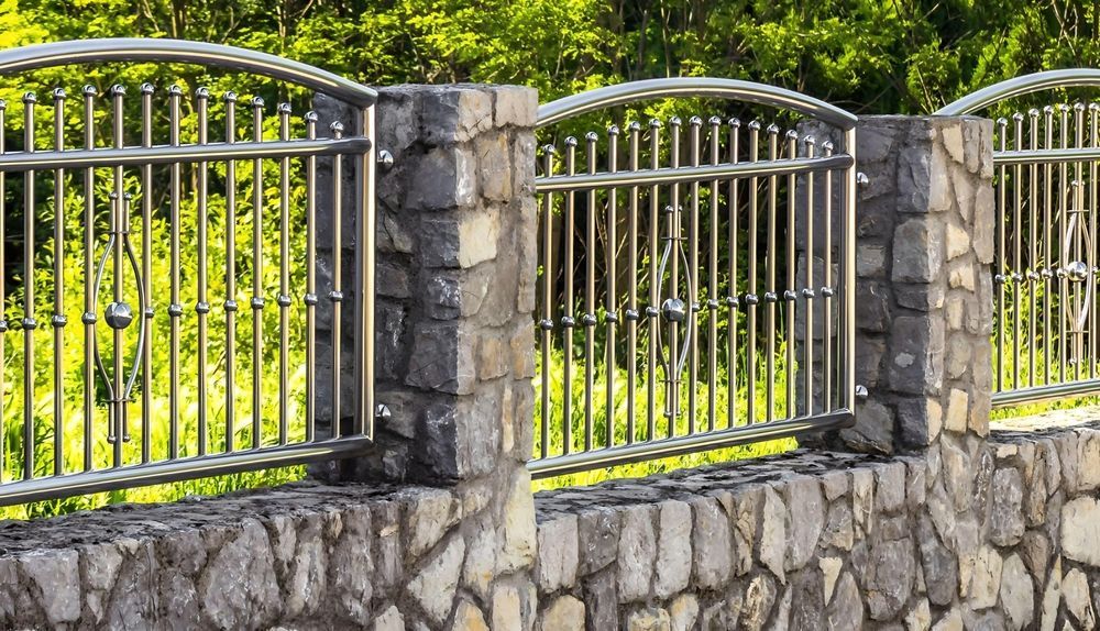 Stone fence with sections of curved, silver-colored metal railings set against a background of green foliage.