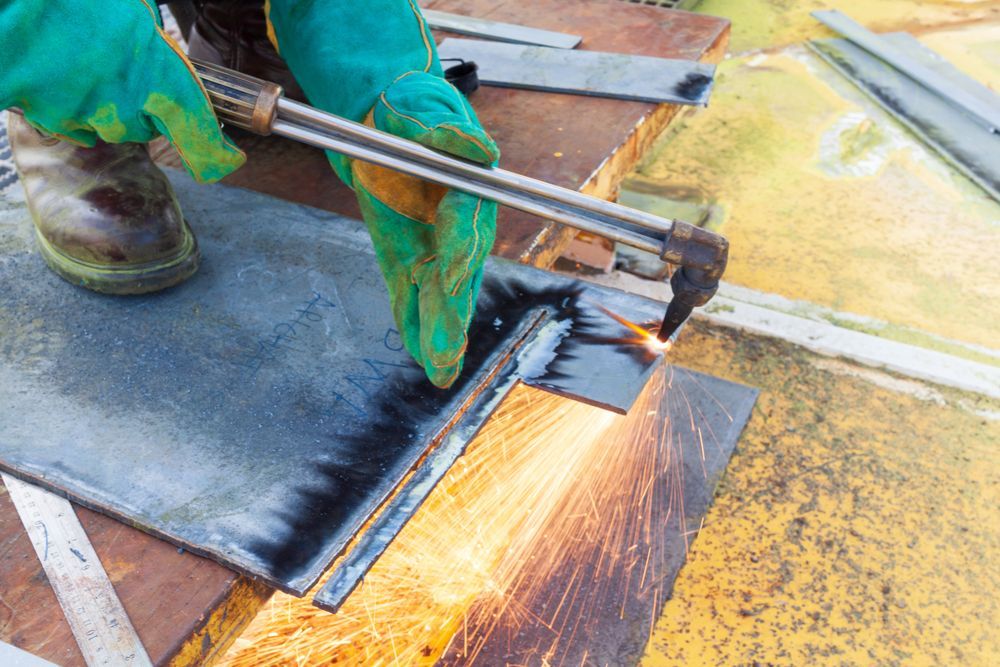 A person wearing protective gloves uses an oxy-fuel torch to cut a thick steel plate on a metal work table.