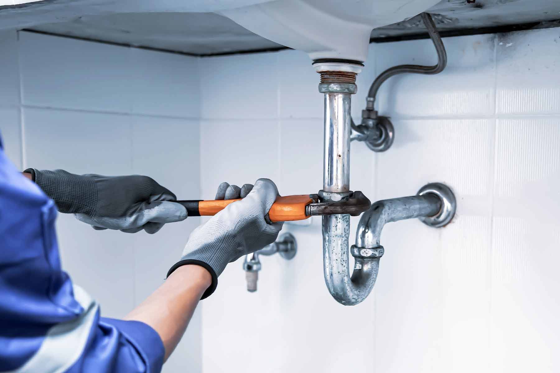 A plumber in a blue uniform uses a wrench to work on the pipes under a white sink, wearing gray gloves. A plumber in a blue uniform uses a wrench to work on the pipes under a white sink, wearing gray gloves.