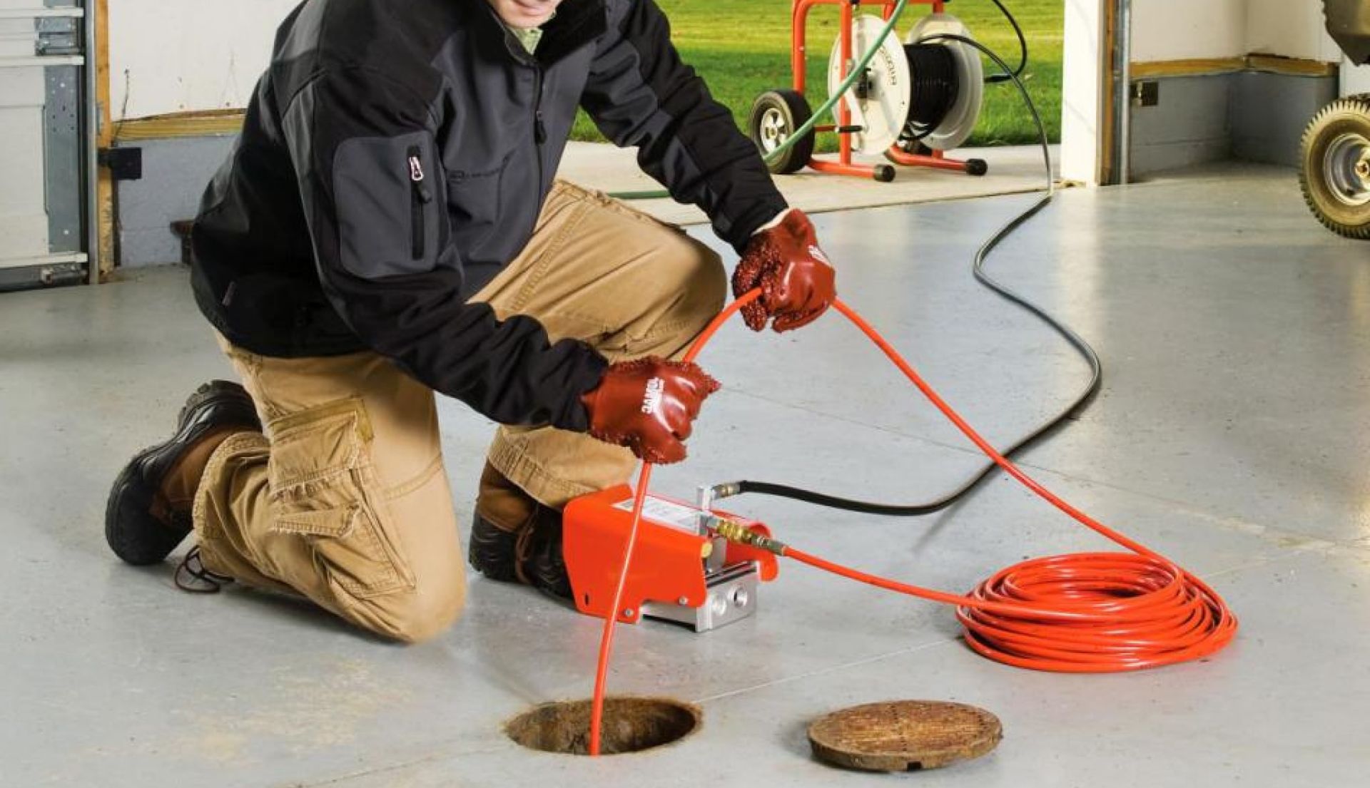 A person in work clothes uses a drain cleaning machine. The setting is a garage, with a coiled orange cable, and a brown drain opening. A person in work clothes uses a drain cleaning machine. The setting is a garage, with a coiled orange cable, and a brown drain opening.
