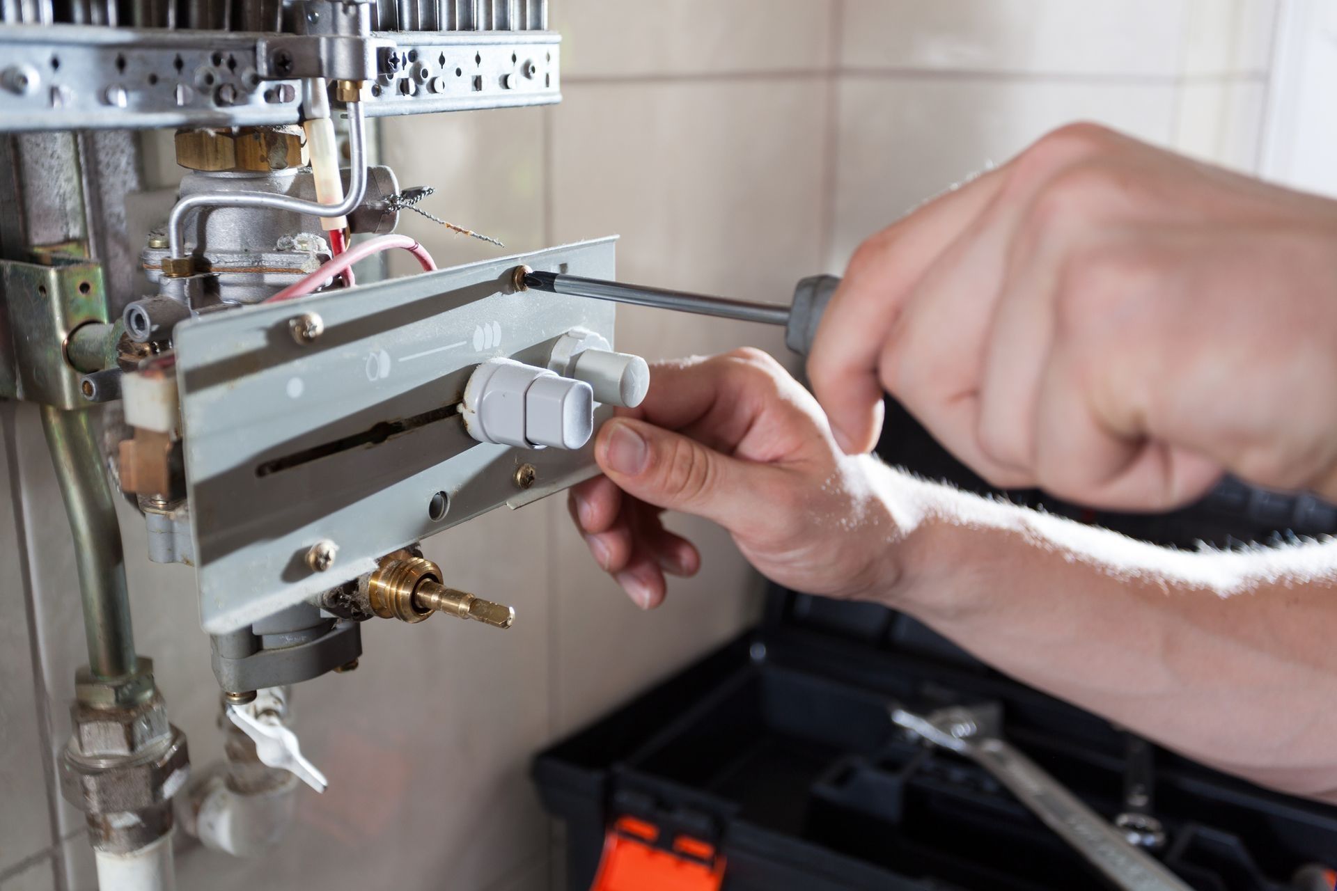 A person uses a screwdriver to work on a water heater. A toolbox sits below. A person uses a screwdriver to work on a water heater. A toolbox sits below.