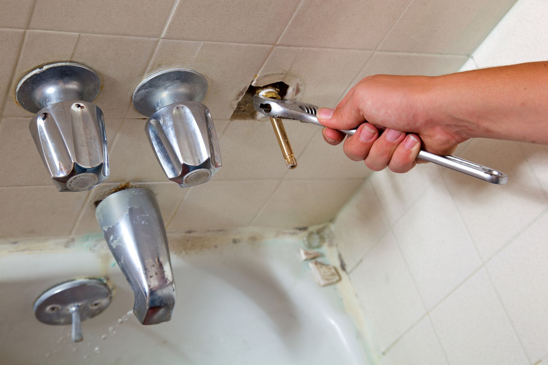 A person using a wrench to fix a bathroom faucet. The faucet is old and silver, with a white-tiled wall.