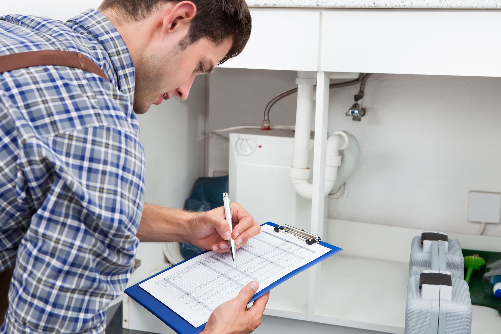 A plumber in a blue plaid shirt inspects pipes under a sink, writing on a clipboard.