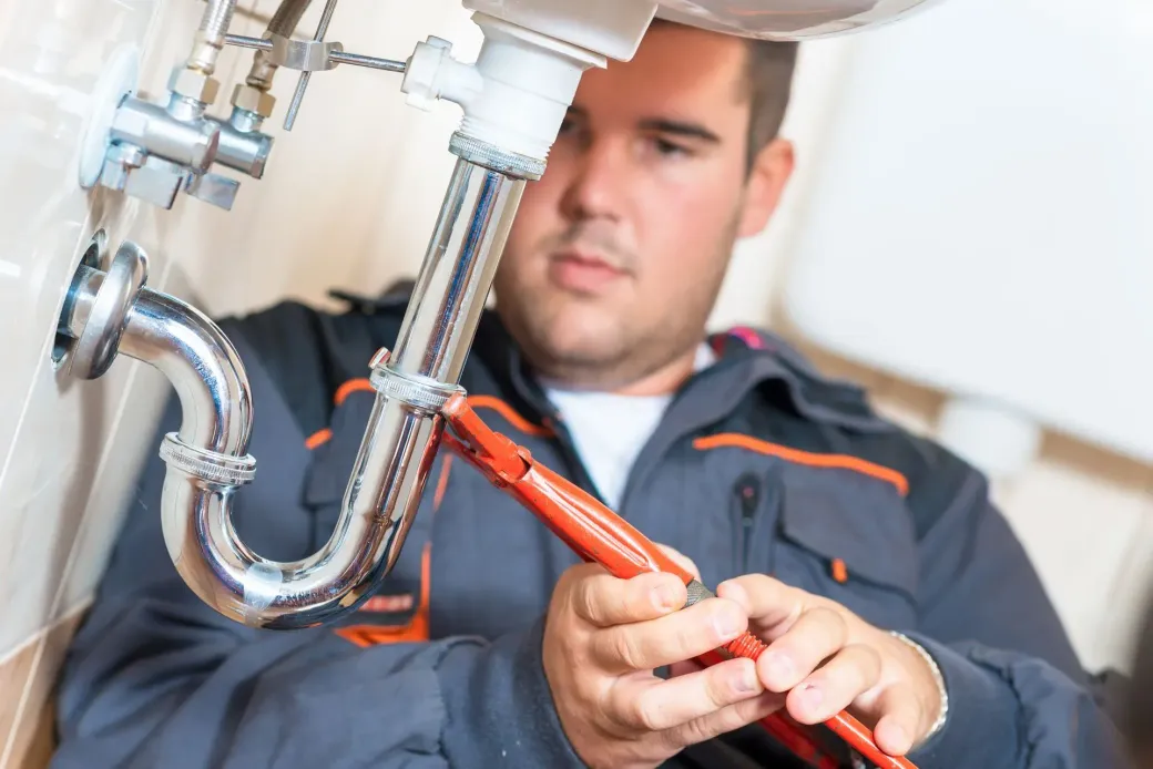 A plumber in a gray jumpsuit uses a wrench to work on a sink's plumbing. He is focused and in a bathroom setting.
