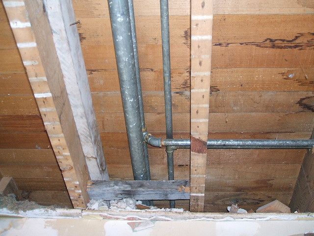 Overhead view inside a building's frame, showing plumbing pipes running through wooden beams and framing. Overhead view inside a building's frame, showing plumbing pipes running through wooden beams and framing.