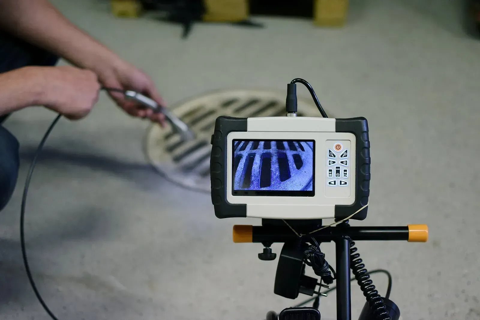 A person using an inspection camera to view a drain grate, the camera's display shows the grate's interior. A person using an inspection camera to view a drain grate, the camera's display shows the grate's interior.