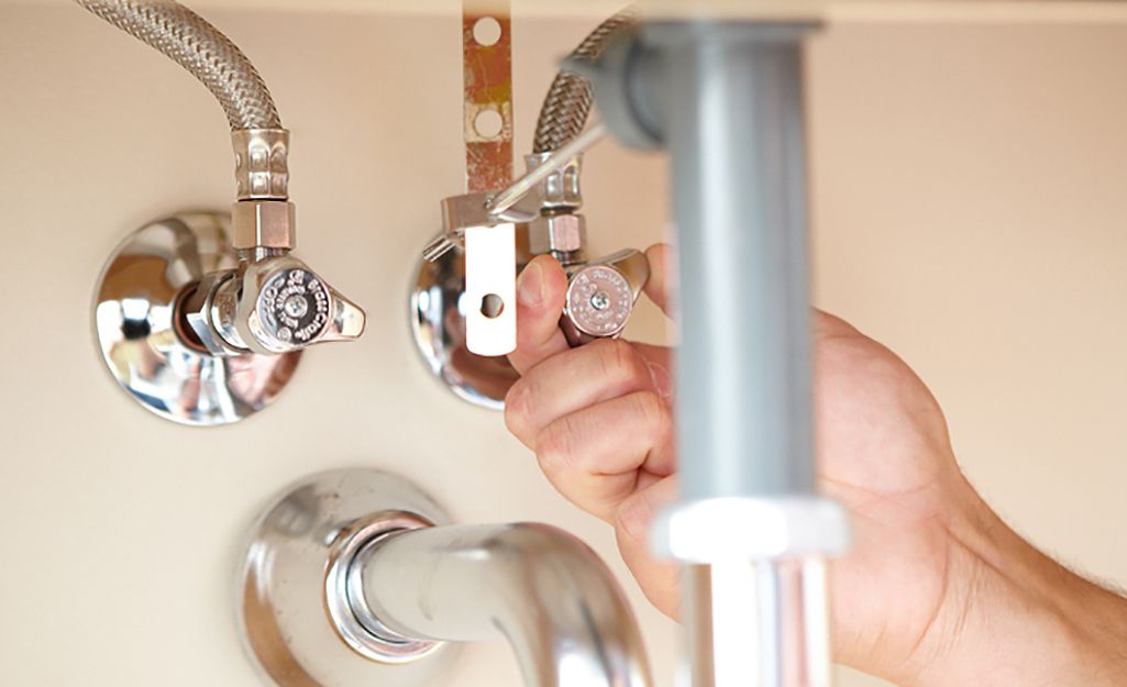 A person's hand turning a water shut-off valve under a sink, near pipes and supply lines.