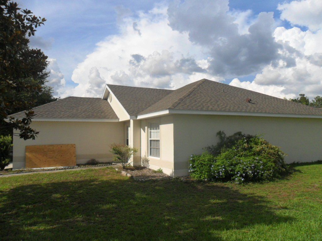 Sunny blue skies with a white house with a gray roof. and big green bush next to the house.