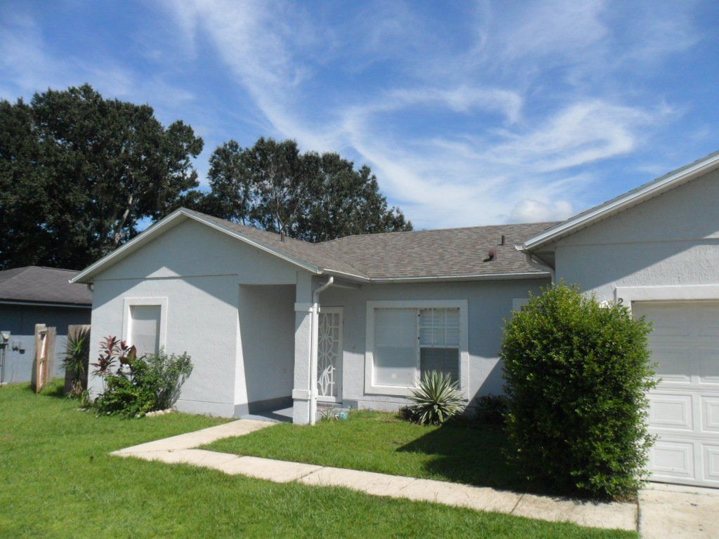 Sunny blue skies, light gray house with a dark gray roof, plants and a bush outside. Small walkway that leads to the front door.
