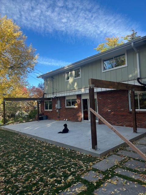 Backyard patio beside a two-story house, with a black dog sitting on the concrete slab.