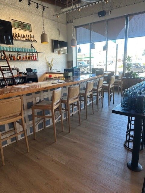 Bright café interior with a long wooden bar, stools, shelves of bottles, and large windows.