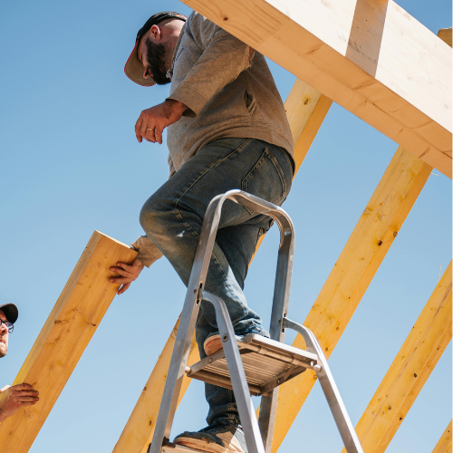 Worker standing on a step ladder framing a wooden roof structure against a blue sky