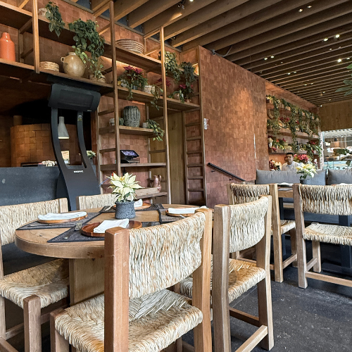 Cozy rustic restaurant interior with wooden tables, woven chairs, shelves, and plants