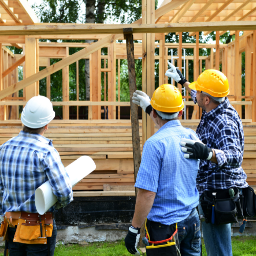 Three construction workers in hard hats inspect wooden house framing at a building site.