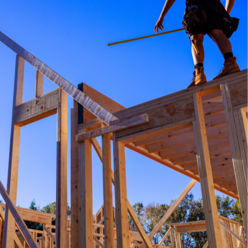 Construction worker on a wooden house frame, standing on the roof under a clear blue sky.