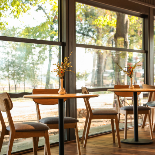 Sunlit café with wooden tables and chairs by large windows overlooking trees