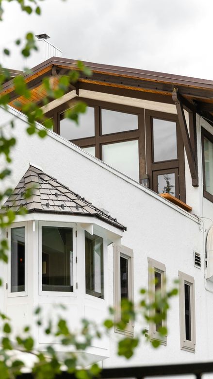 White house with brown timber accents and a small bay window, viewed through blurred green leaves.