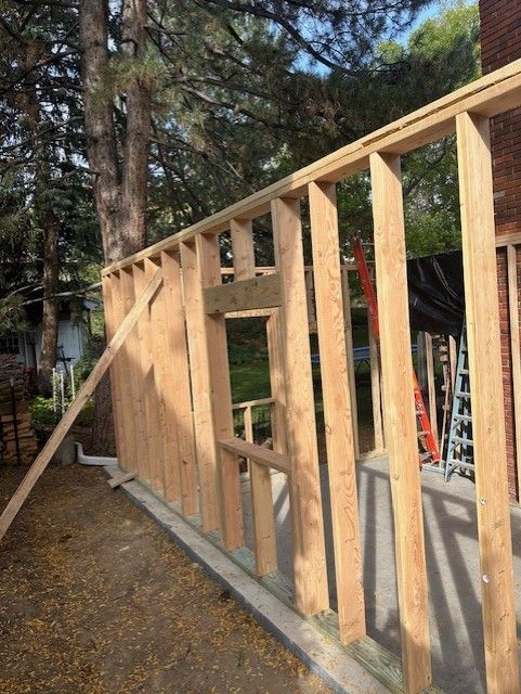 Wood-framed wall under construction beside a wooded area, with open window and door openings
