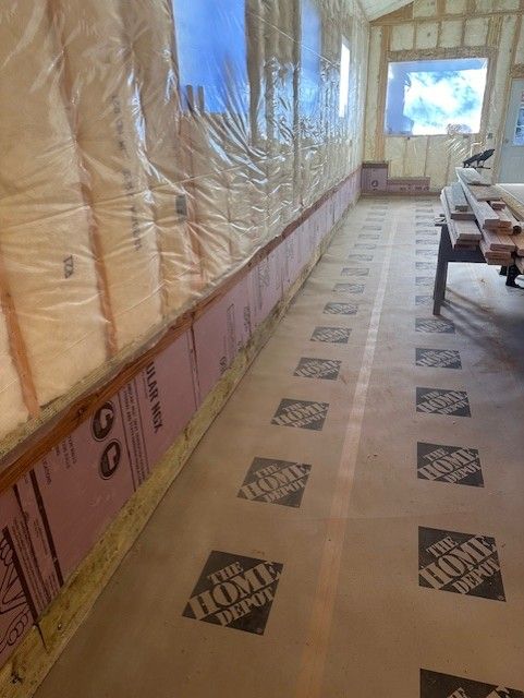 Insulated hallway under construction with protected flooring, taped walls, and a bright window at the end.