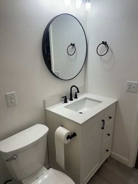 Small white bathroom with toilet, vanity sink, round mirror, and towel rings on the wall.