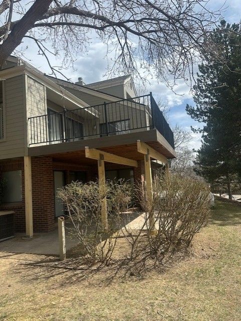 Two-story beige house with balcony, brick pillars, and leafless shrubs in a yard