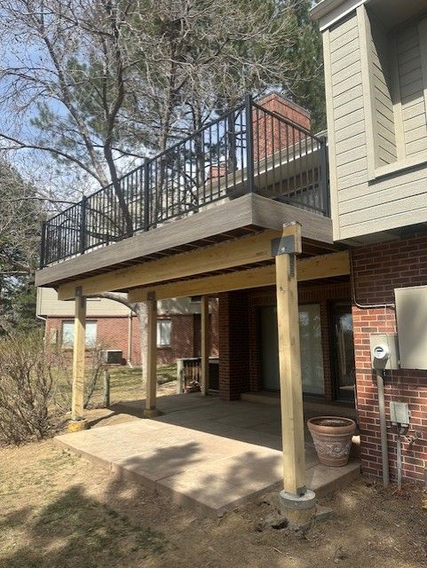 Covered patio beneath a brick house with black railing balcony and concrete slab steps