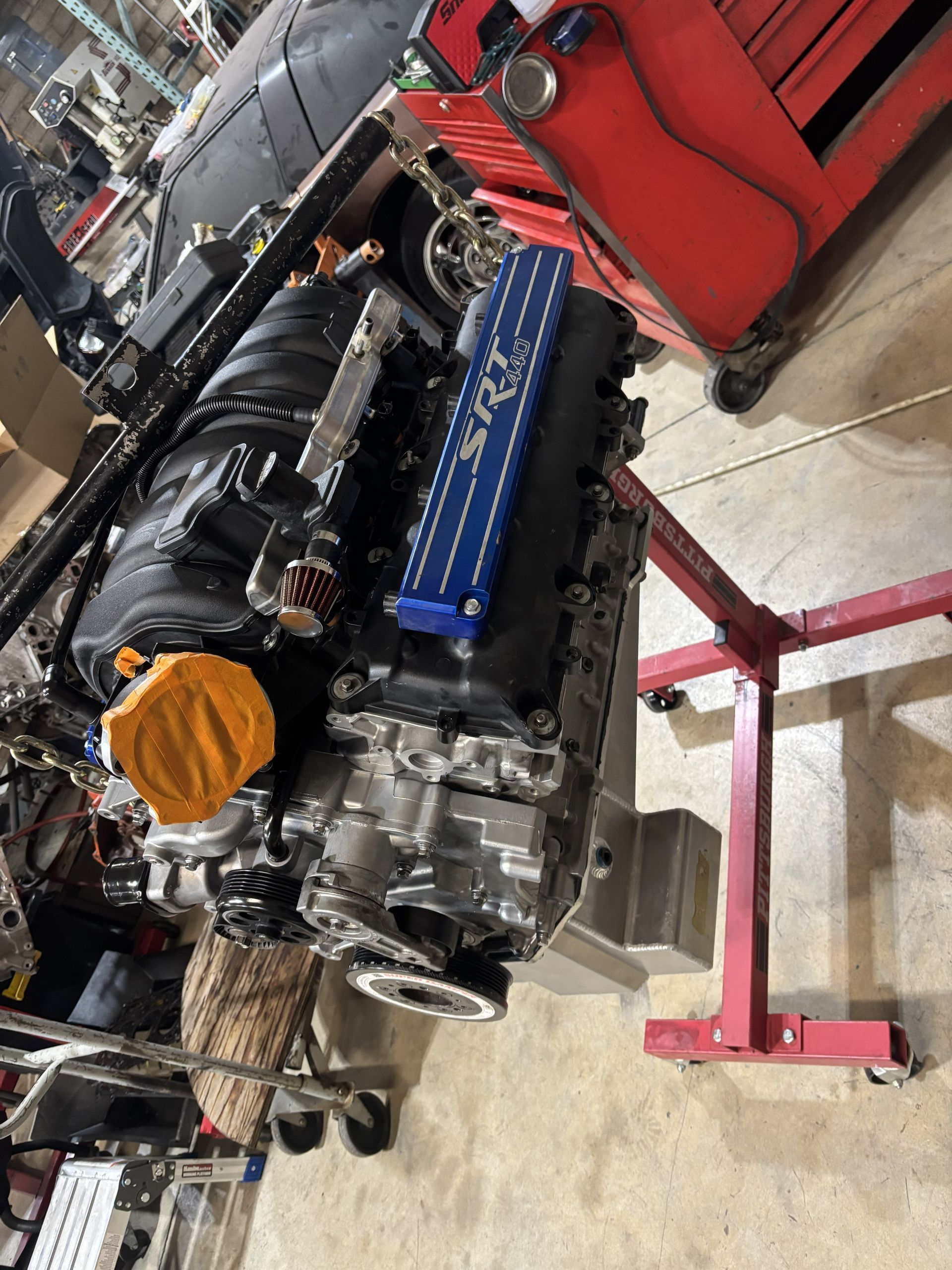 Engine on a red stand; blue valve cover, black components, orange oil cap, garage setting.