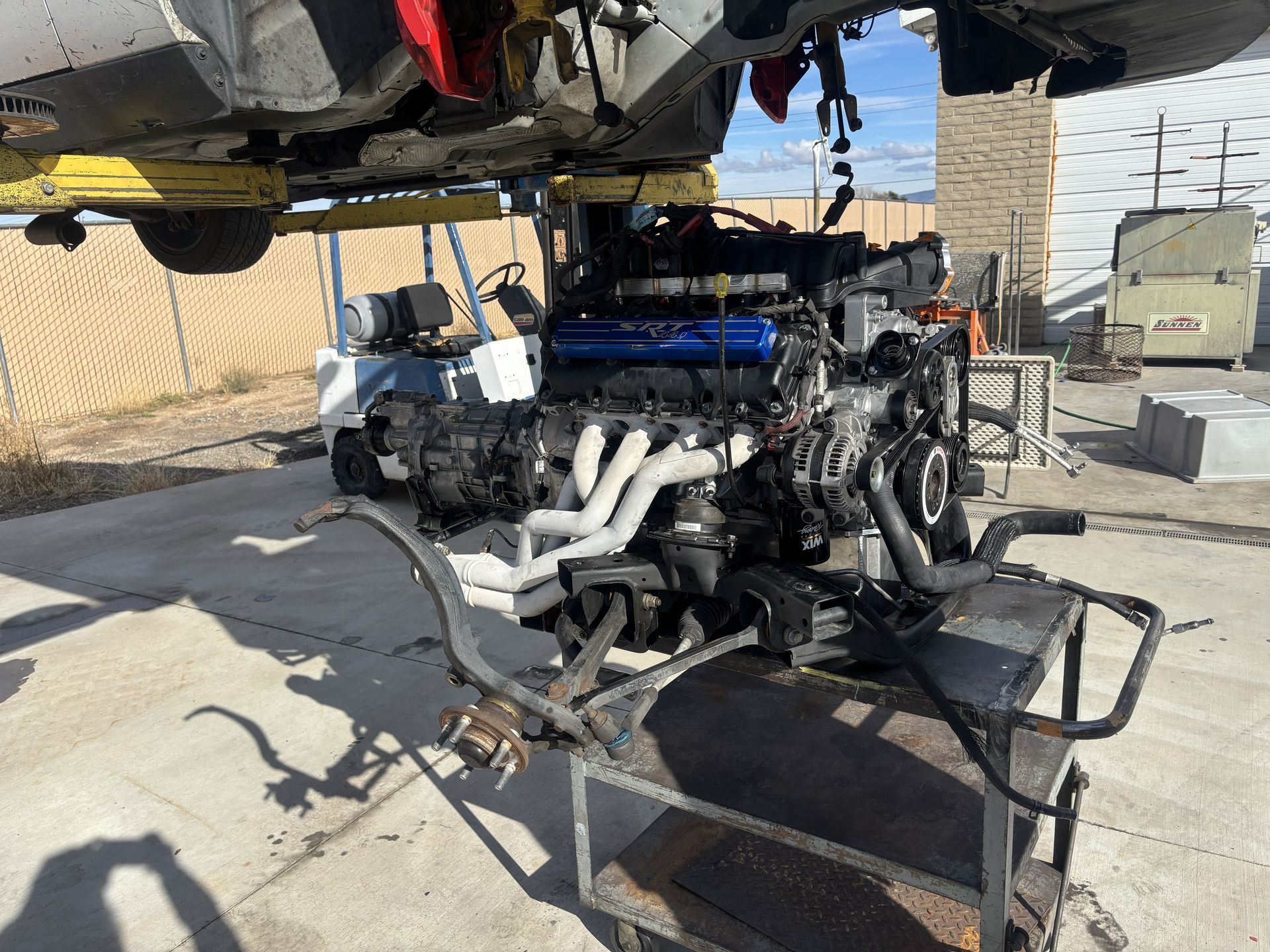 Car engine on cart, beneath a vehicle on a lift outdoors. Engine has white exhaust pipes and blue valve cover.