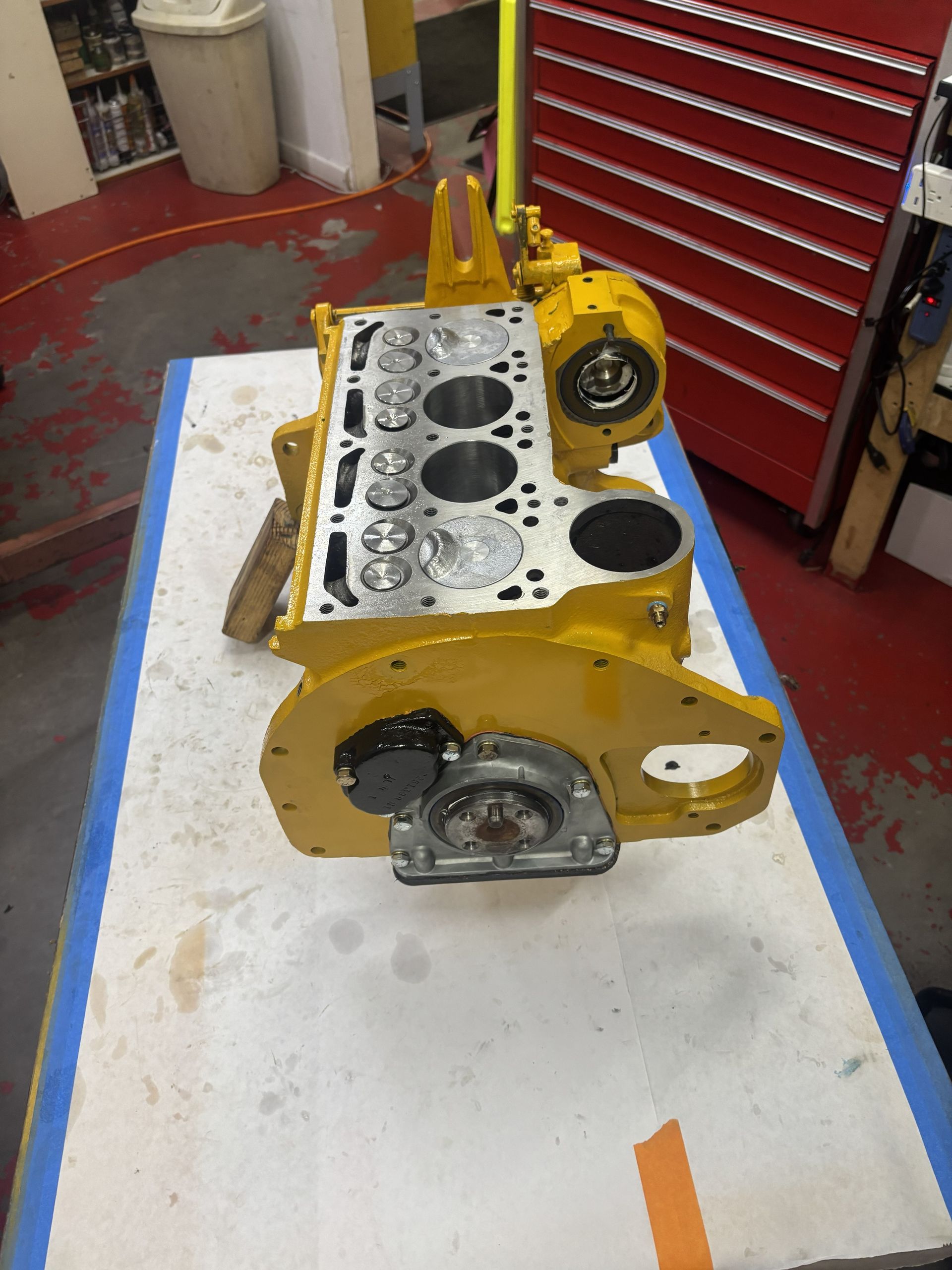 Yellow engine block on a workbench, possibly undergoing repair, with exposed cylinders and associated components.