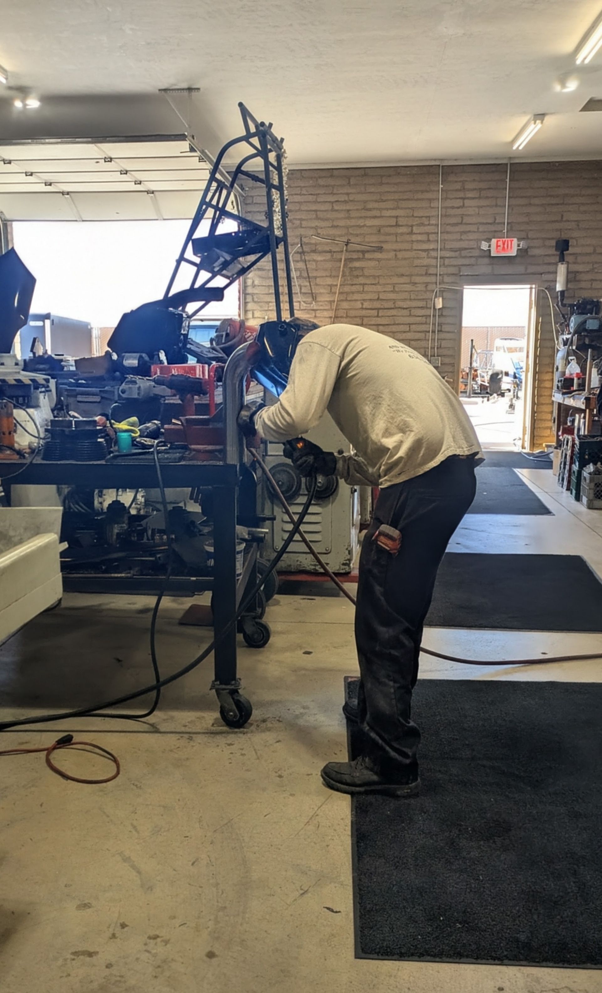 A person welding metal in a workshop, wearing protective gear. They are leaning over a workbench near tools.