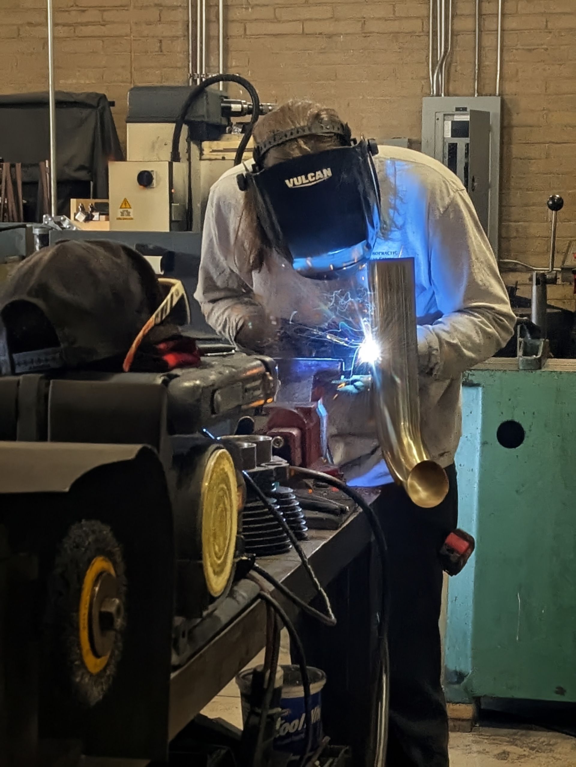 Welder in a workshop wearing a mask, welding a gold-colored pipe, sparks flying.