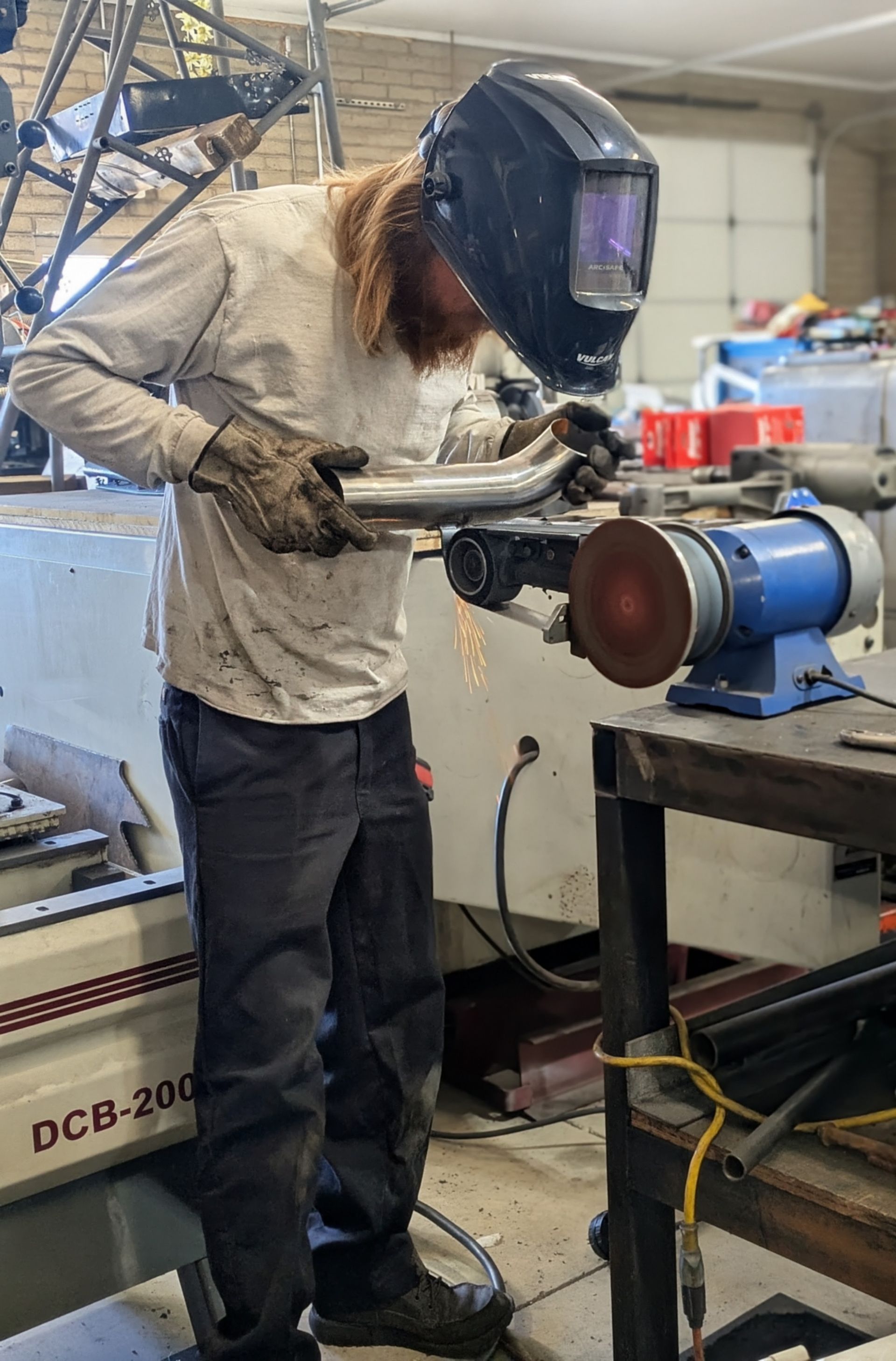 Person welding metal tube while wearing a mask and gloves, using a grinder in a workshop.