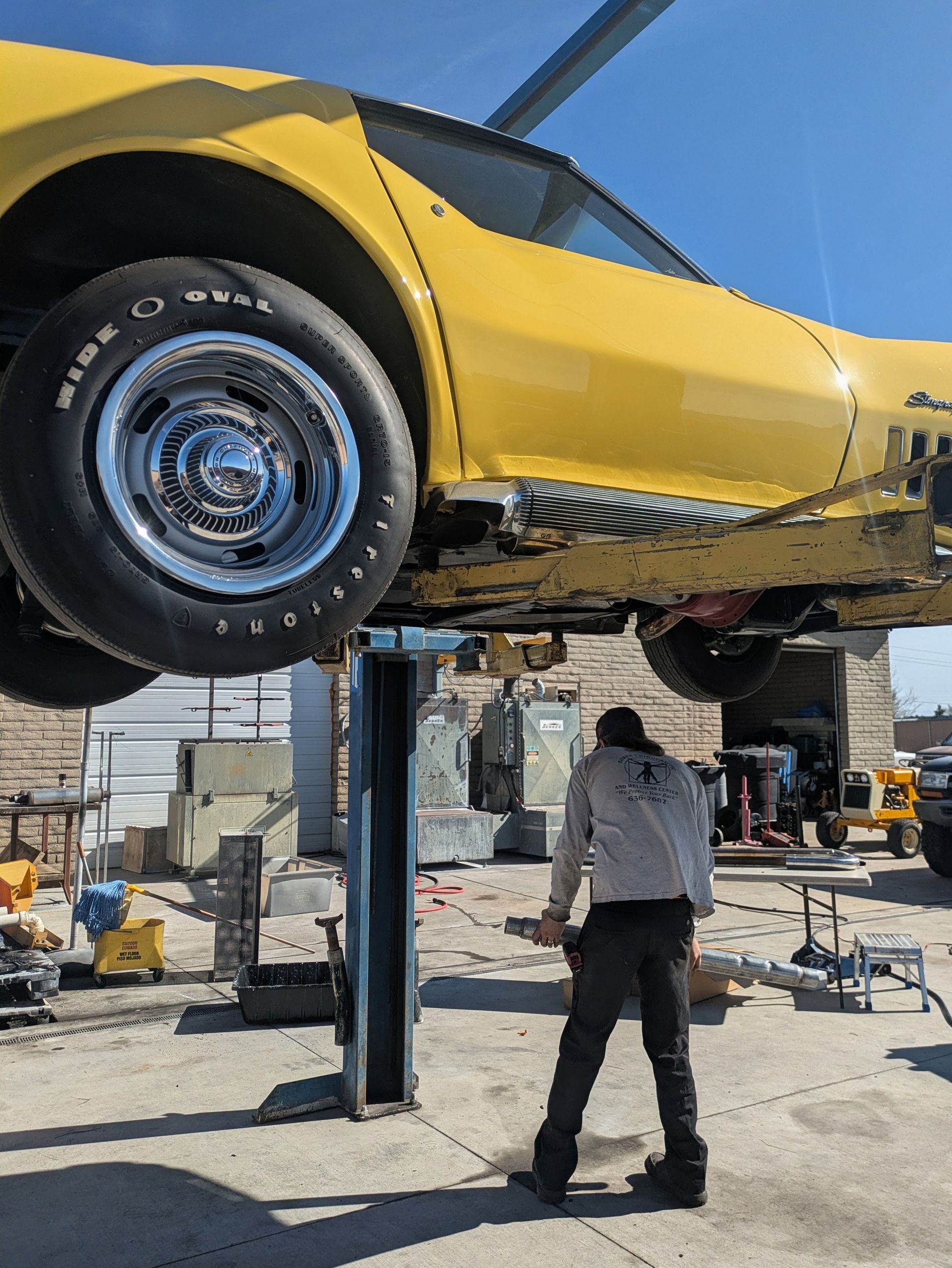 Yellow Corvette on a lift in a shop, mechanic working beneath it on a sunny day.