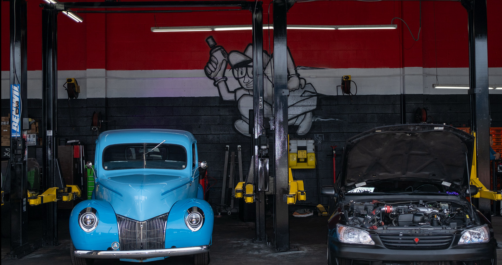 Blue vintage truck and black car in a garage with a lift, black and red walls, and a cartoon mural.