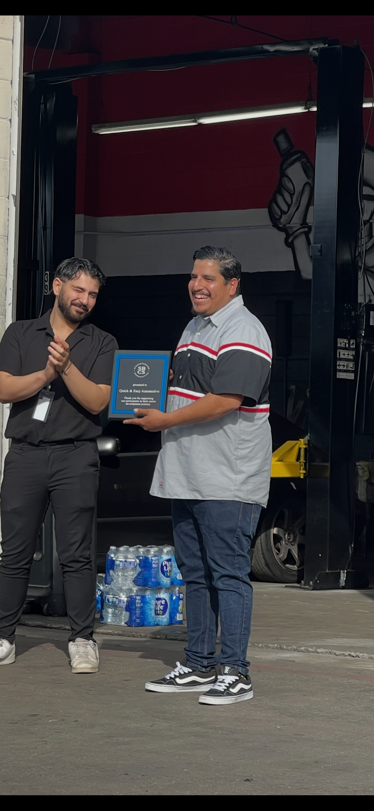 Two men in an auto shop, one holding an award plaque, the other clapping.