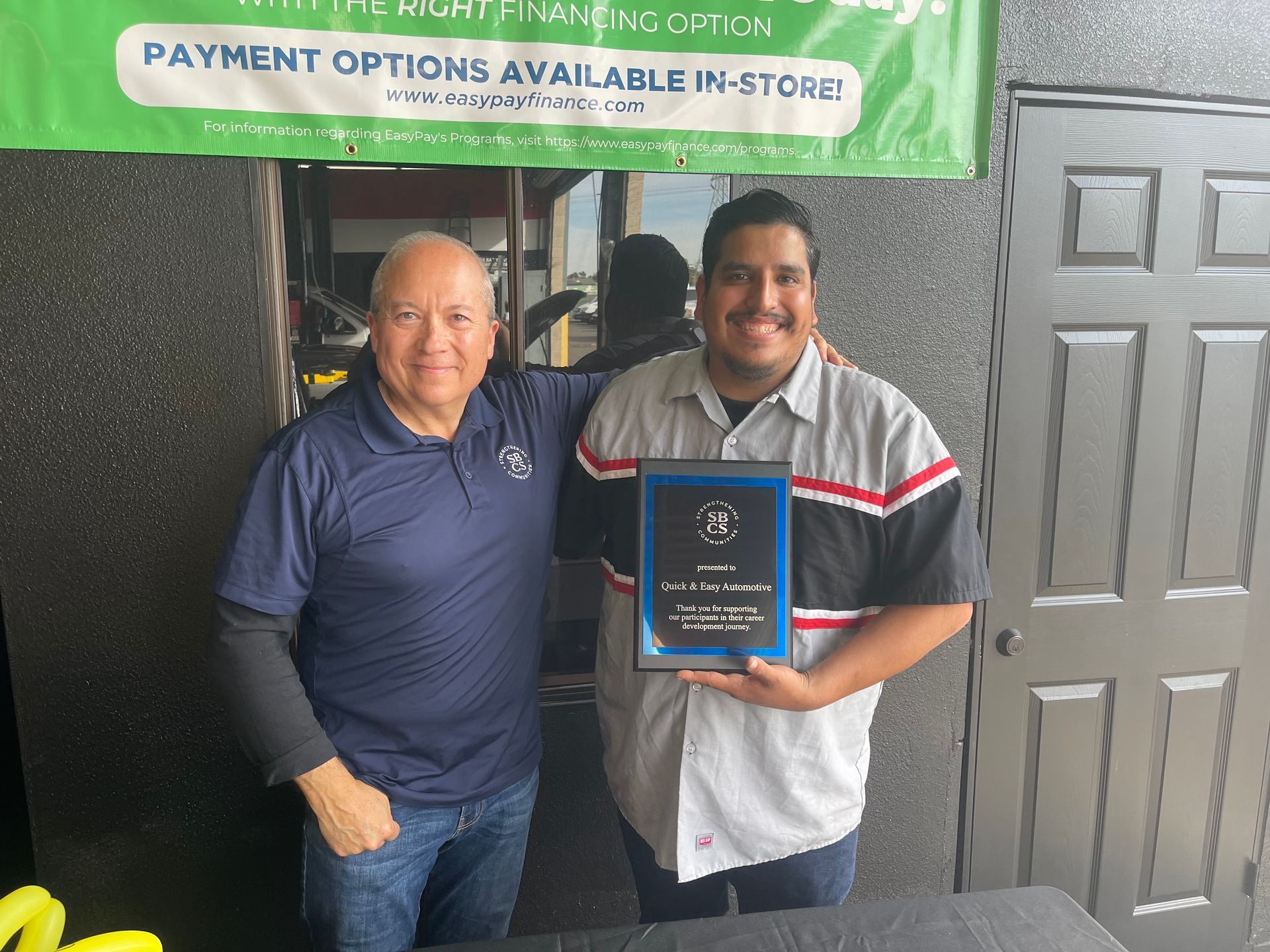 Two men pose, one holding a plaque.  They are smiling, standing near a building with a banner above.
