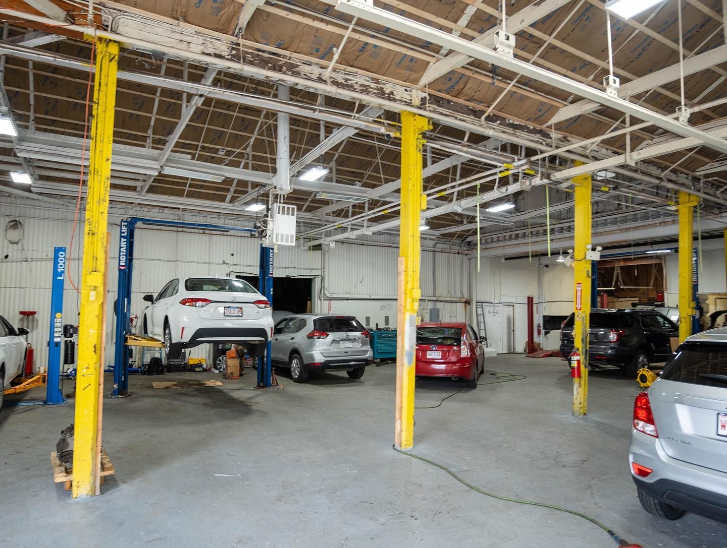 Inside a car repair shop with vehicles on lifts and scattered around. Yellow support beams and a gray floor. | JR Car Care Inc