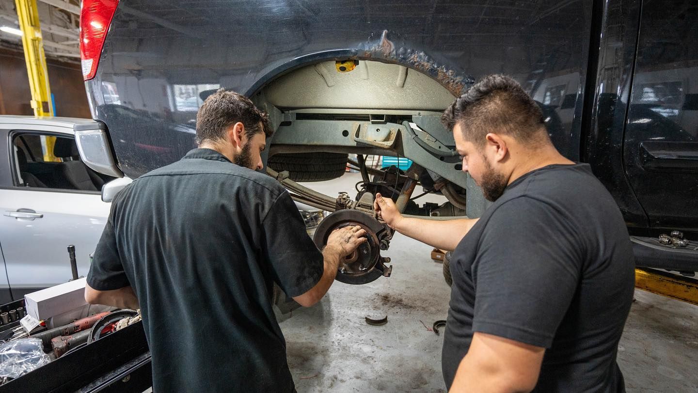 Two mechanics working on vehicle's wheel in a repair shop. One points to parts, the other works. | JR Car Care Inc