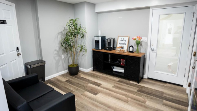 Office waiting area with a black sofa, console with coffee maker, and plant. Door and light-colored wood floor. | JR Car Care Inc