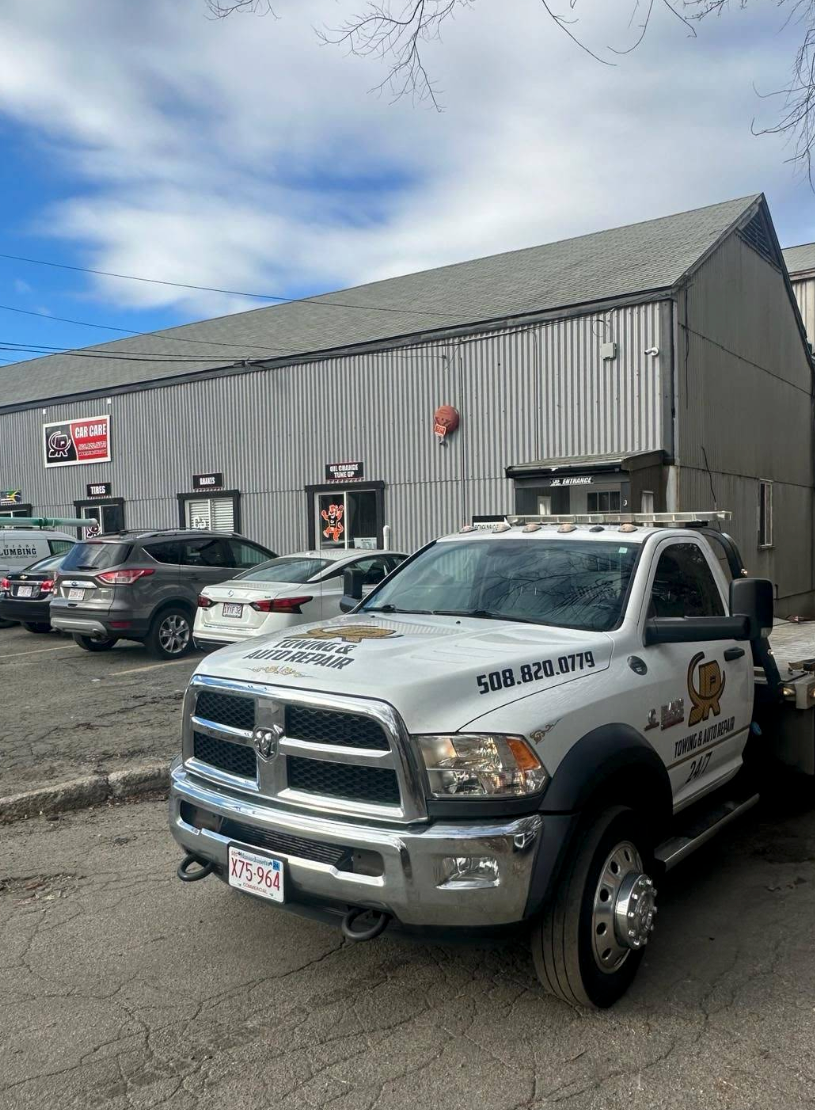 Tow truck parked in front of a building with cars. White and gold truck has company logo. Gray building, cloudy sky. | JR Car Care Inc