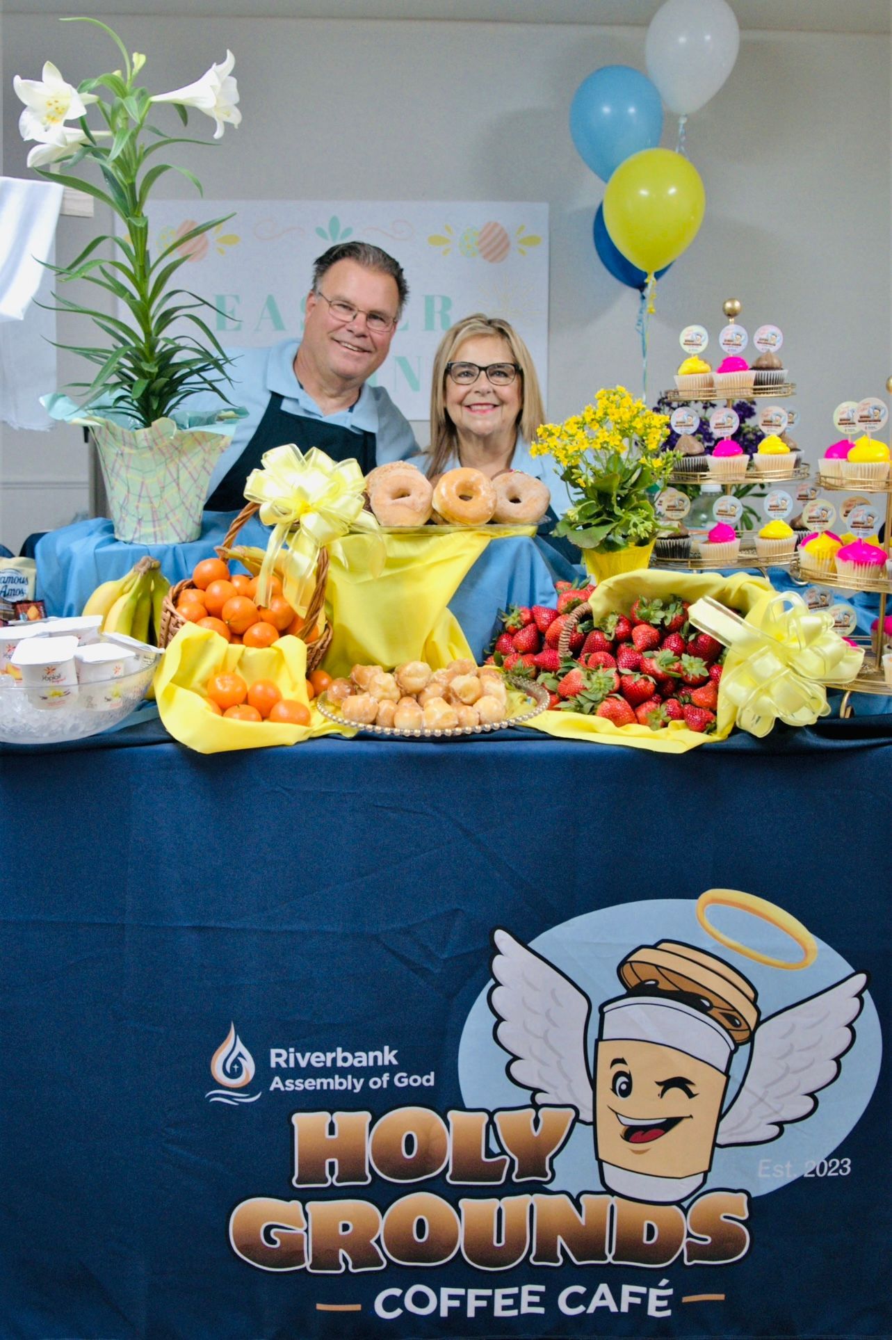 A man and a woman are standing behind a table with a sign that says holy grounds coffee cafe.
